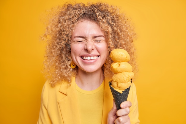 Free photo joyful positive curly haired woman closes eyes from happiness smiles broadly holds tasty appetizing ice cream of lemon flavor in waffle expresses sincere authentic emotions. monochrome shot.