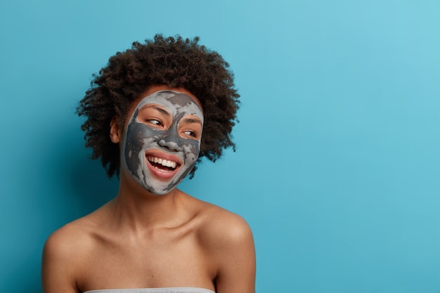 Joyful positive dark skinned curly woman stands naked indoor, applies beauty mud mask for perfect soft facial skin, cares about complexion, looks gladfully away, isolated on blue wall.