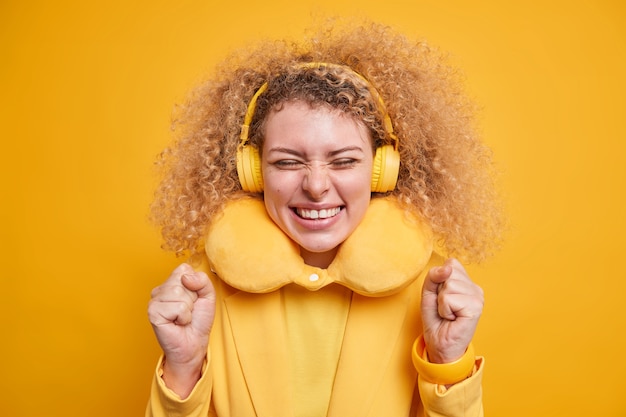 Free photo joyful positive woman with curly hair wears comfortable pillow around neck to reduce pain enjoys favorite music uses wireless headphones expresses happiness isolated over yellow  wall