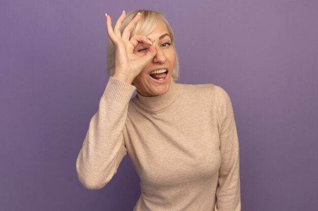 Joyful pretty blonde slavic woman looks at camera through fingers on purple