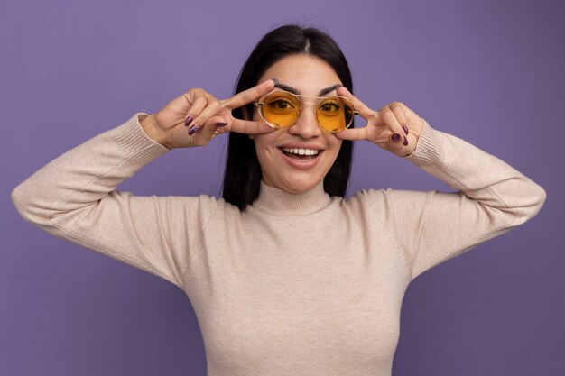 Joyful pretty brunette woman in sun glasses gestures victory hand sign with two hands isolated on purple wall