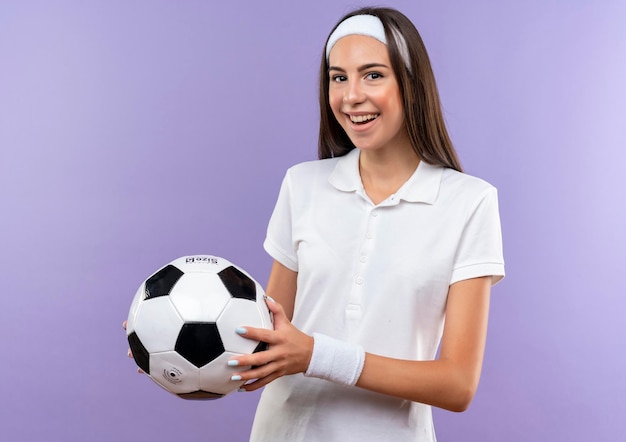 Joyful pretty sporty girl wearing headband and wristband holding soccer ball isolated on purple wall