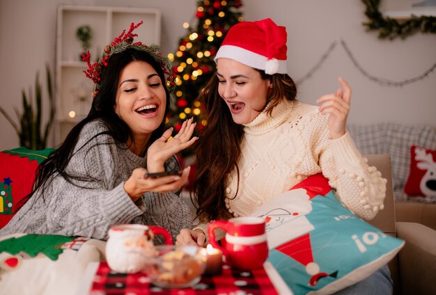 Joyful pretty young girls with santa hat look at phone sitting on armchairs and enjoying christmas time at home