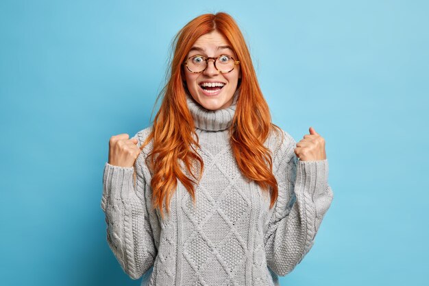 Joyful redhead young woman with cheerful expression rejoices success clenches fists after achievement goals dressed in knitted grey sweater.