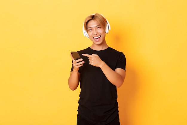 Joyful satisfied asian handsome guy, listening music or good podcast in headphones, pointing finger at smartphone with pleased smile, yellow wall