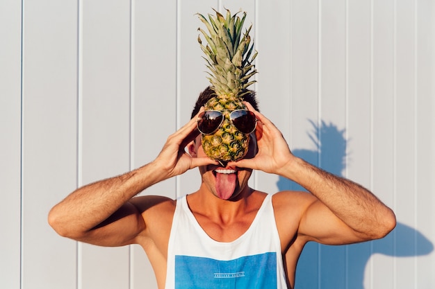 Free photo joyful smiling man showing a tongue, holding a pineapple with sunglasses