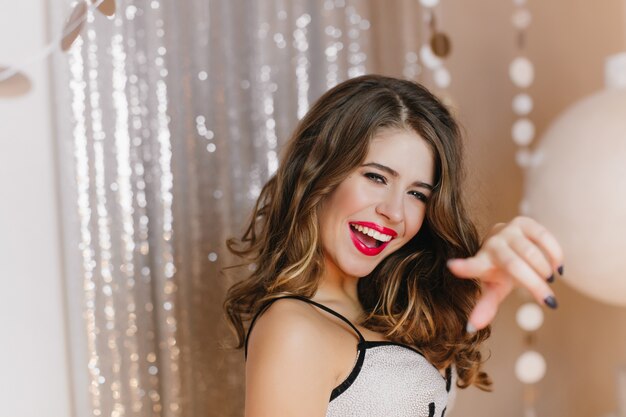 Joyful, spectacular dark-haired girl slyly looks and point with her finger. Photo of woman posing on shiny wall