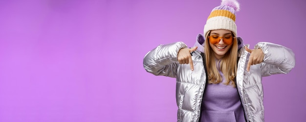 Free photo joyful stylish blond woman in silver jacket stylish sunglasses jat enjoying perfect mountains view snowy vacation resort look pointing down smiling amused having fun feel happy purple background