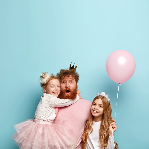 Free photo joyful surprised father forgets to buy some attributes for holiday, spends free time with two cute daughters, celebrate special occasion, have balloon party, isolated over blue wall. fathers day