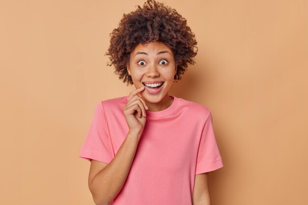 Joyful surprised woman smiles broadly keeps finger near mouth feels amazed dressed in casual pink t shirt hears something awesome gets surprise isolated over brown background. Happy emotions