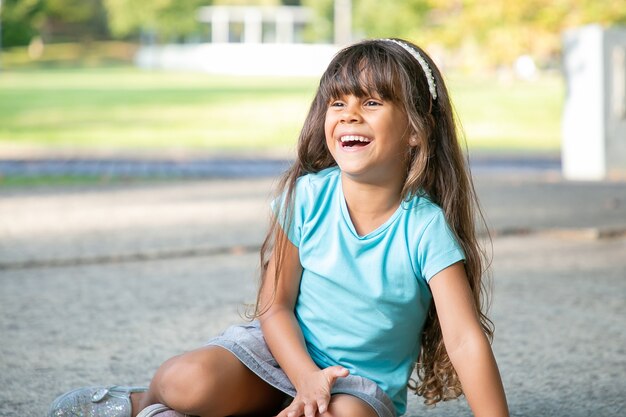 Joyful sweet black haired girl sitting on ground, looking away and laughing. Childhood and outdoor activity concept