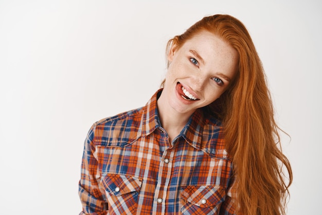 Joyful teenage girl with red hair and pale skin showing tongue and smiling happy at camera standing over white background