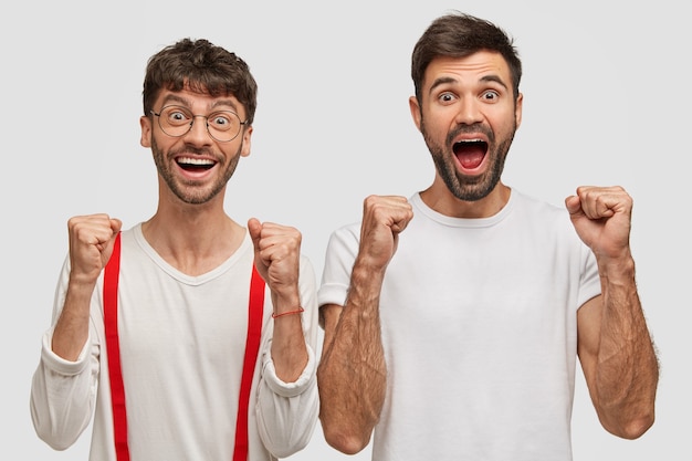 Joyful unshaven two young men clench fists and shout with happiness, dressed casually, isolated on white wall