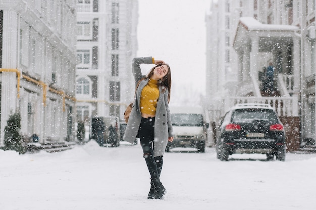 Joyful winter young woman chilling in snowfall on street in big city. Fashionable model with backpack, smiling with closed eyes, enjoying snowing, snowflakes, christmas mood.