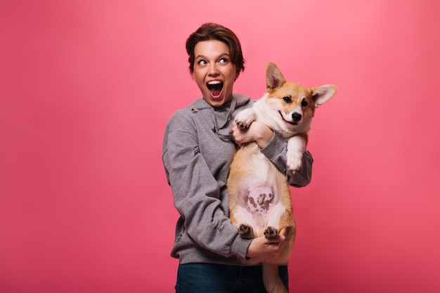 Joyful woman in grey hoodie holding corgi on pink background. Emotional teen girl in jeans plays with dog on isolated