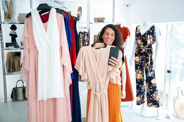 Joyful woman shopping in clothes store and consulting friend on cellphone, showing dress on hanger. Boutique customer or communication concept