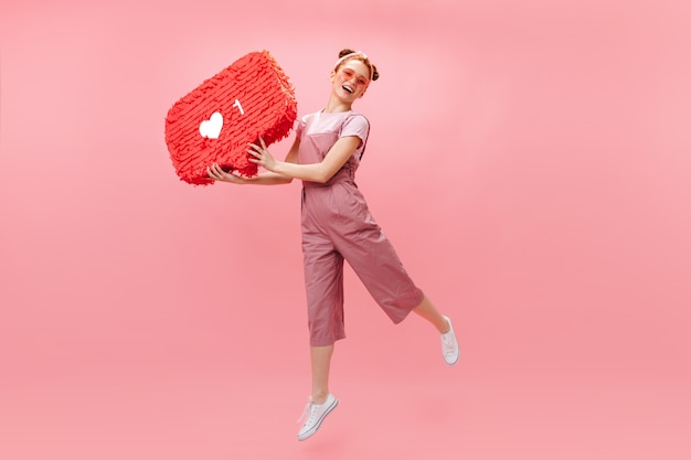 Free Photo joyful woman in stylish jumpsuit jumping on pink background, holding like sign.