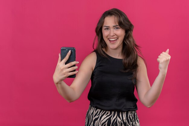 Joyful woman wearing black undershirt listen music from phone and dancing on pink wall