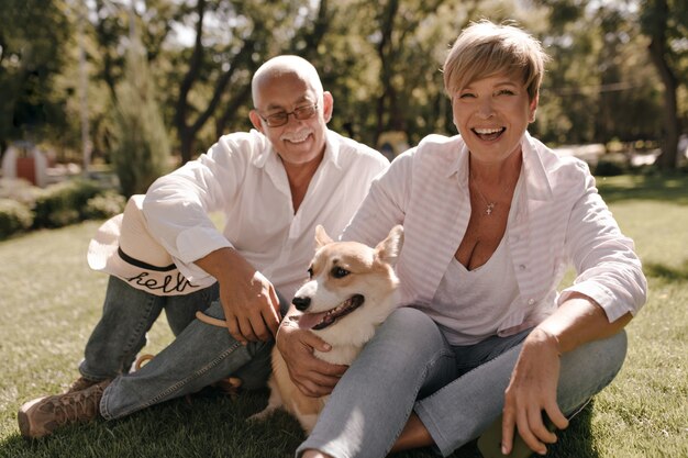 Joyful woman with blonde trendy hairstyle in pink shirt and jeans laughing, hugging corgi and sitting with grey haired man in eyeglasses in park.