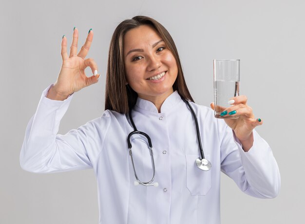 Joyful young asian female doctor wearing medical robe and stethoscope holding glass of water looking at front doing ok sign isolated on white wall