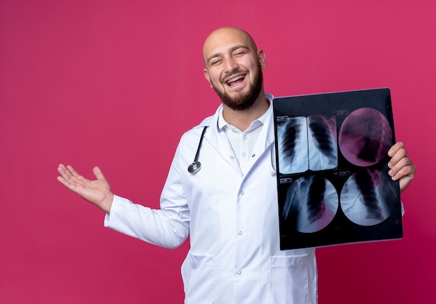 Free photo joyful young bald male doctor wearing medical robe and stethoscope holding x-ray and spread hand