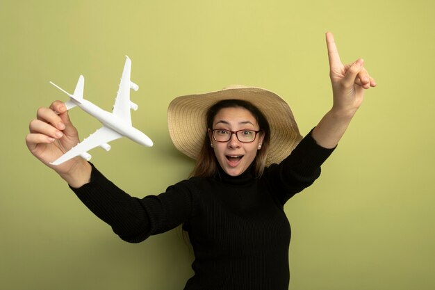 Joyful young beautiful girl in a black turtleneck and glasses holding toy airplane lookign at camera surprised showing index finger haing great idea 