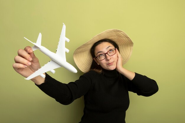 Joyful young beautiful girl in a black turtleneck and glasses holding toy airplane looking up thinking positive 