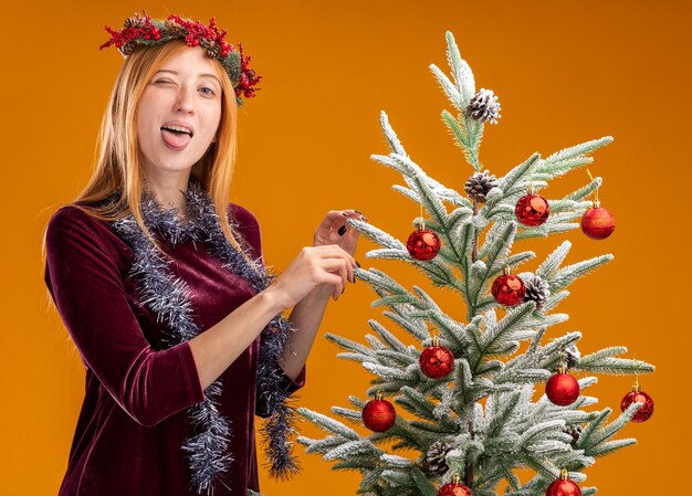 Joyful young beautiful girl standing nearby christmas tree wearing red dress and wreath with garland on neck showing tongue isolated on orange wall