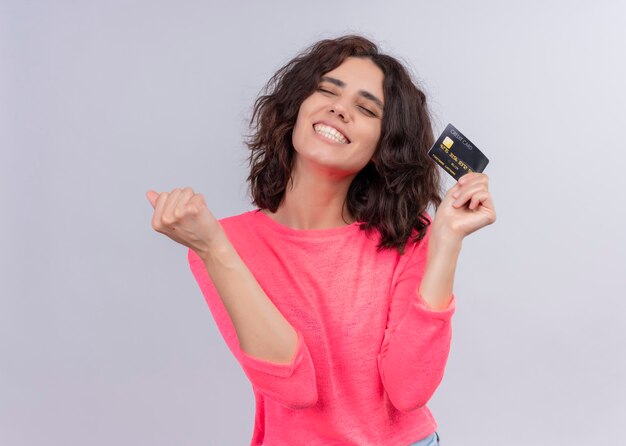 Joyful young beautiful woman holding card and raising fist on isolated white wall