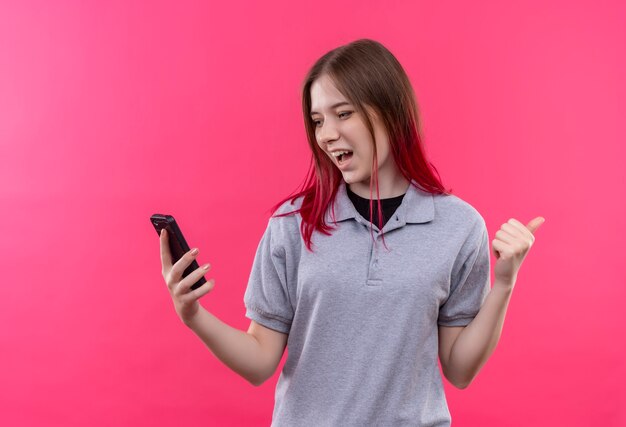 Joyful young beautiful woman wearing gray t-shirt lookinh at phone in her hand showing yes gesture on isolated pink wall