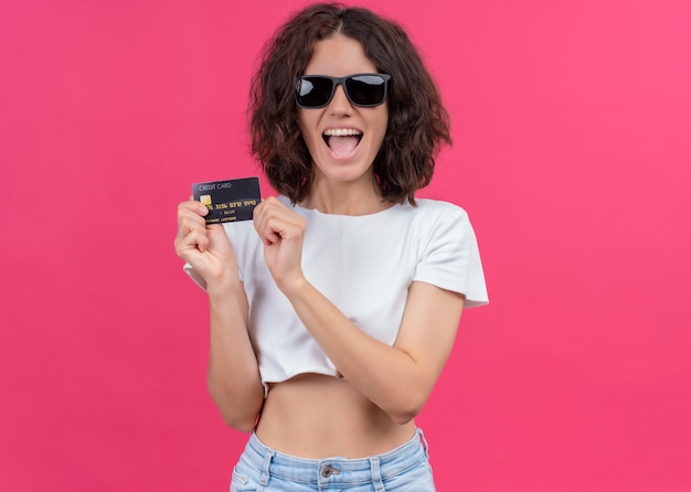 Joyful young beautiful woman wearing sunglasses and holding card on isolated pink wall with copy space