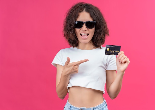 Joyful young beautiful woman wearing sunglasses and holding card pointing at it on isolated pink wall