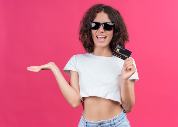 Joyful young beautiful woman wearing sunglasses and holding card showing empty hand on isolated pink wall