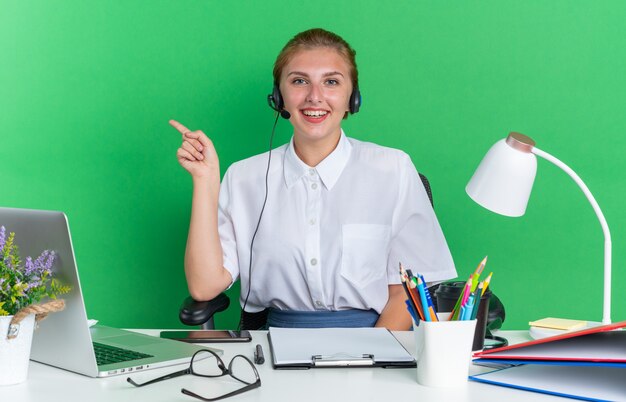 Joyful young blonde call centre girl wearing headset sitting at desk with work tools looking at camera pointing at side isolated on green wall