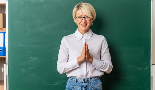 Free photo joyful young blonde female teacher wearing glasses in classroom standing in front of chalkboard keeping hands together looking at camera