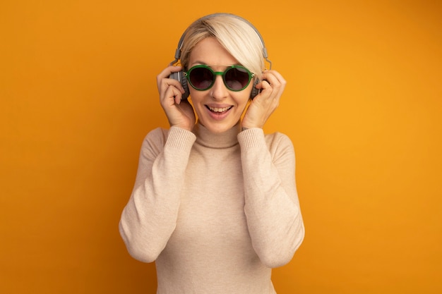 Free photo joyful young blonde girl wearing sunglasses and headphones grabbing headphones listening to music isolated on orange wall with copy space