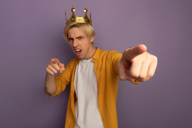 Joyful young blonde guy wearing yellow t-shirt and crown showing you gesture isolated on purple