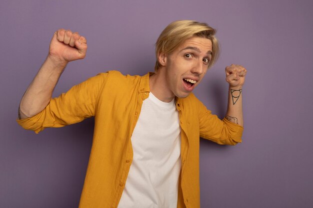 Joyful young blonde guy wearing yellow t-shirt raising hands isolated on purple with copy space