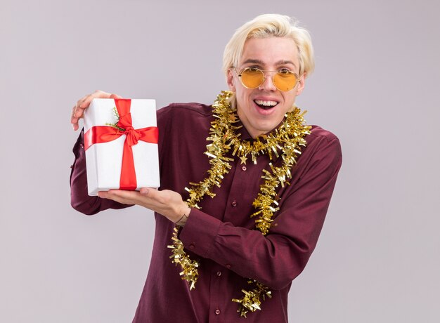 Joyful young blonde man wearing glasses with tinsel garland around neck holding gift package looking at camera isolated on white background