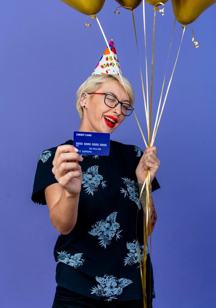 Joyful young blonde party woman wearing glasses and birthday cap holding balloons and credit card winking at front isolated on purple wall