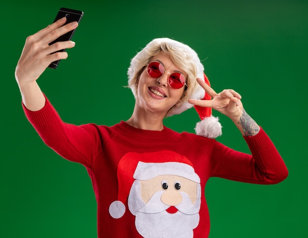 Free photo joyful young blonde woman wearing christmas hat and santa claus christmas sweater with glasses doing peace sign taking selfie isolated on green background