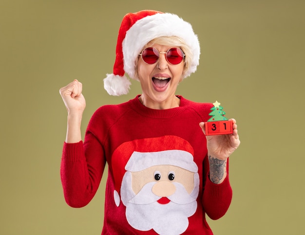 Free photo joyful young blonde woman wearing christmas hat and santa claus christmas sweater with glasses holding christmas tree toy with date doing yes gesture with closed eyes isolated on olive green wall