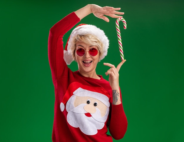 Free Photo joyful young blonde woman wearing christmas hat and santa claus christmas sweater with glasses looking at camera holding christmas candy cane near head isolated on green background