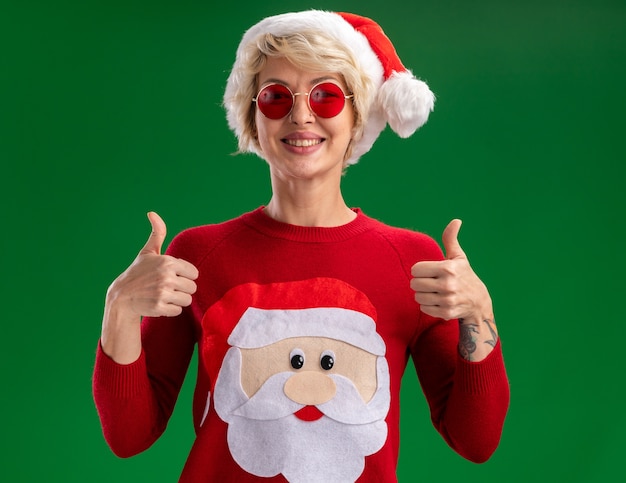 Free Photo joyful young blonde woman wearing christmas hat and santa claus christmas sweater with glasses looking at camera showing thumbs up isolated on green background