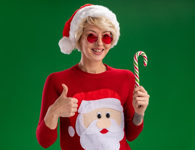 Joyful young blonde woman wearing christmas hat and santa claus christmas sweater with glasses looking  holding christmas candy cane showing thumb up isolated on green wall