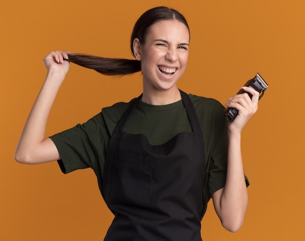 Joyful young brunette barber girl in uniform holds her braid and hair clippers isolated on orange wall with copy space