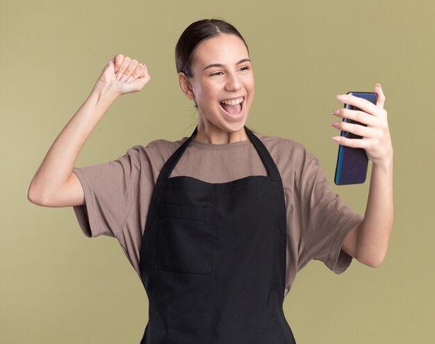 Joyful young brunette barber girl in uniform keeps fist holding and looking at phone on olive green