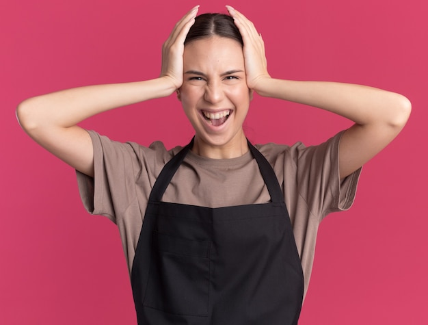 Joyful young brunette barber girl in uniform puts hands on head isolated on pink wall with copy space