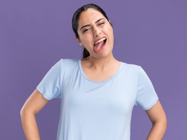 Joyful young brunette caucasian girl blinks eye and stucks out tongue isolated on purple wall with copy space