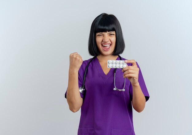 Joyful young brunette female doctor in uniform with stethoscope keeps fist and holds medicine 